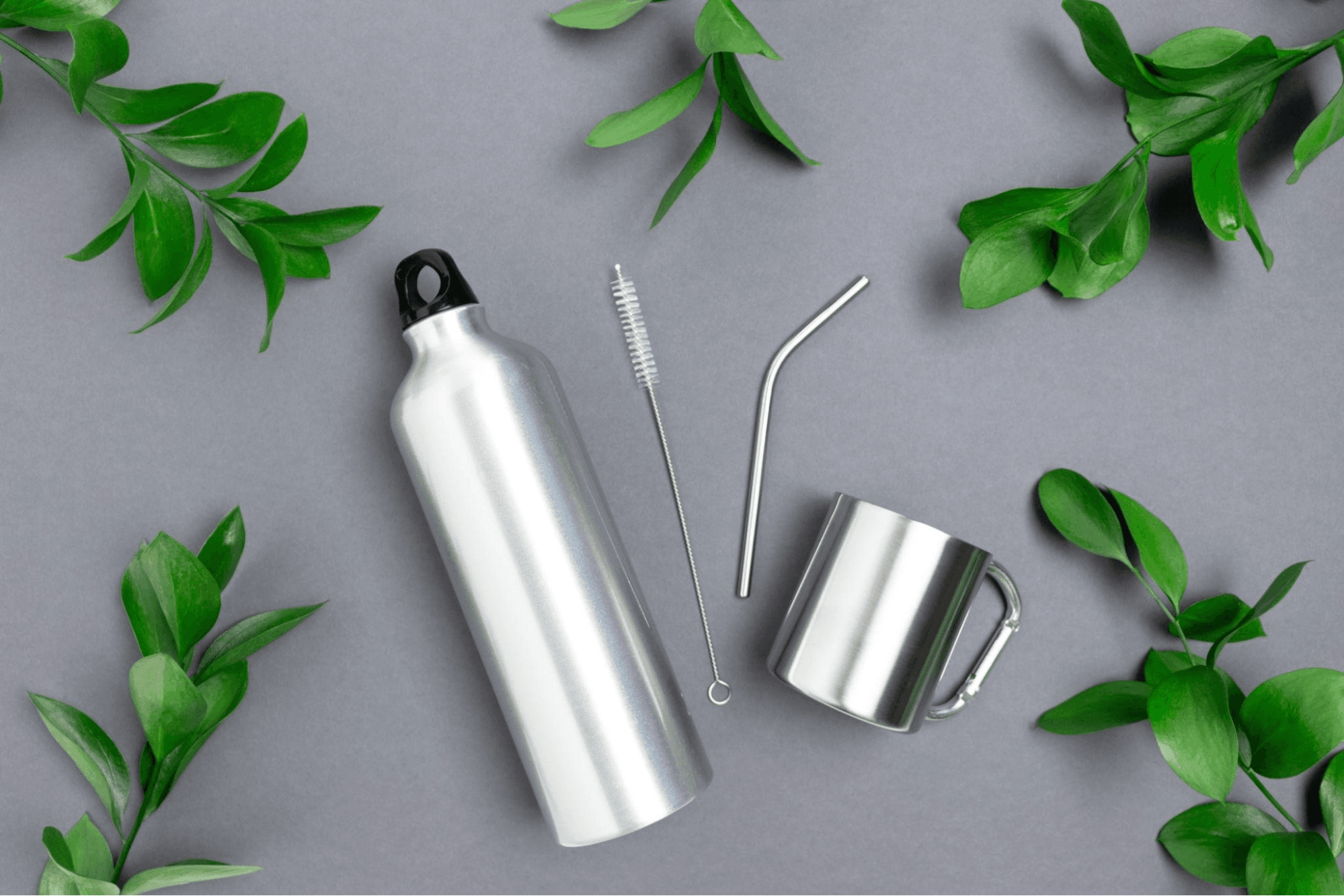 Aluminium bottle, cleaning brush, bent metal straw and steel camping mug arranged among green leaves